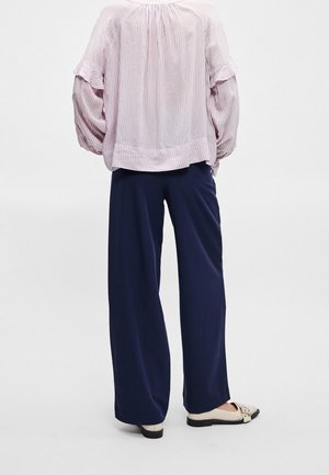 Personne portant un pantalon ample bleu marine, des chaussures crème sans lacets et un chemisier à rayures rose clair à manches longues bouffantes, debout devant un fond uni.