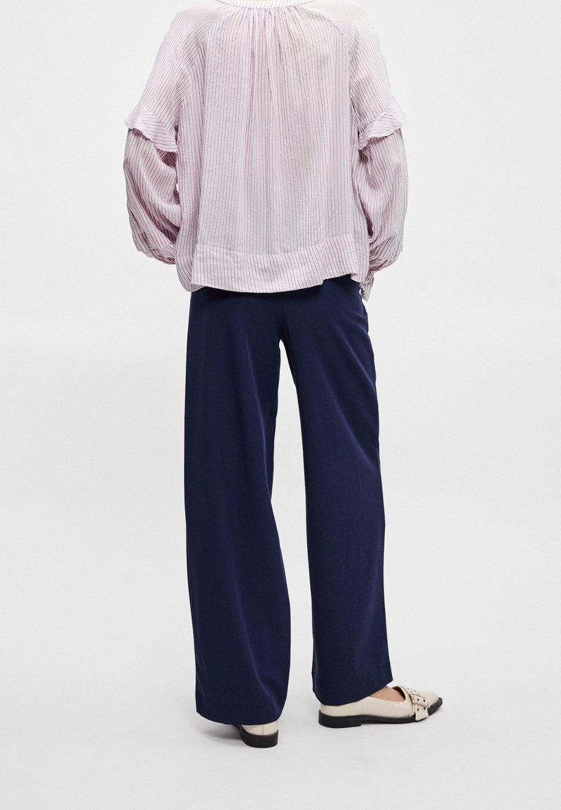 Personne portant un pantalon ample bleu marine, des chaussures crème sans lacets et un chemisier à rayures rose clair à manches longues bouffantes, debout devant un fond uni.