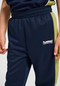 Granatowe sportowe spodnie z elastycznym paskiem; posiadają limonkowo-zielone boczne panele oraz biały napis „hummel PERFORMANCE”. Gładki, lekki materiał.