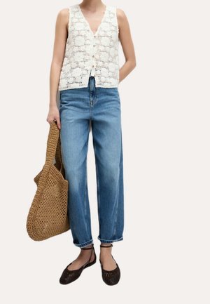 Femme portant un gilet en crochet blanc sans manches, un jean bleu taille haute, des ballerines tissées noires, et tenant un grand sac fourre-tout tissé marron.