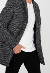 Kabát z vlněné směsi v černobílém houndstooth vzoru, s jednímřadým zapínáním, s klopami a postranními kapsami, navlečený přes bílou košili.