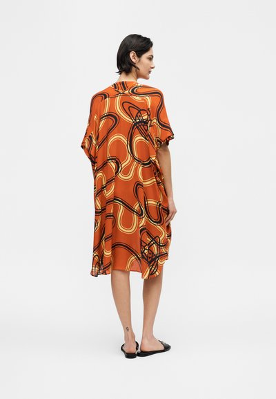 Γυναίκα με κοντά σκούρα μαλλιά που φοράει πορτοκαλί φόρεμα με μαύρες και κίτρινες αφηρημένες στροφές, όρθια ξυπόλητη στραμμένη ελαφρώς αριστερά σε λευκό φόντο.