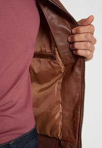 Bruine leren jas open gedragen om een gladde, satijnachtige voering met een klein zakje te tonen; gedragen over een bordeauxrood shirt.