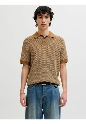 Tânăr cu păr creț închis la culoare, purtând un tricou polo maro texturat cu mâneci scurte și blugi albaștri, stând pe un fundal alb simplu.