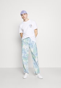 Marmor-mønstrede jeans i pastelfarver som blå og grøn, parret med en hvid T-shirt med et cirkulært logo, og en lyse lilla hue. Hvide sneakers.