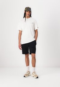Hvit t-skjorte med logo, svarte shorts og beige joggesko. Modellen har på seg en flerfarget camo-lue, hvite sokker og står foran en ensfarget bakgrunn.