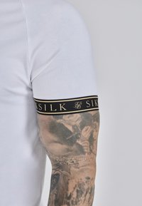 Balts īsās piedurknēs T-krekls ar melnu joslu pie piedurknes, kurā ir zeltīta "SILK" uzraksts. Audums izskatās gluds un piegulošs.