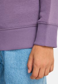 Sweat-shirt violet avec poignets côtelés, présentant une texture douce. Associé à un denim bleu clair, montrant un motif de tissu détaillé.