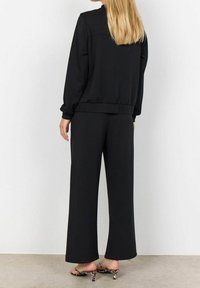 Blonde vrouw in losse zwarte broek, bijpassend zwart lang mouwtop en leopard-print lage instappers, staande op een betonnen vloer.