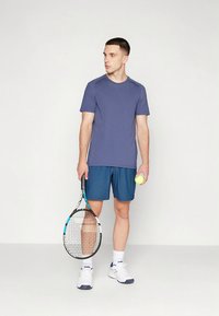 Mannlig modell iført en tettsittende marineblå t-skjorte og blå treningsshorts, som holder en tennisracket og en gul tennisball. Hvite joggesko.