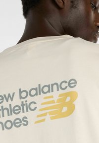 Crème-kleurig T-shirt met een ronde hals, met daarop "new balance sport schoenen" in het grijs en een geel logo op de achterkant.