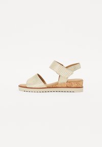 Wedge sandals with a textured gold strap, cork midsole, and white rubber outsole featuring a zigzag pattern. Open toe design.