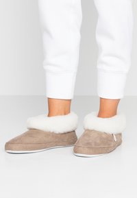Soft beige suede slippers with a plush white fur cuff, rounded toe design, and white rubber sole. Simple stitching detail and side tag visible.