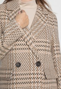 Cappotto doppiopetto in un motivo houndstooth, con colori beige, marrone e bianco. Presenta rever a scollatura e bottoni neri. Tessuto testurizzato.