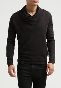 Man som bär en svart långärmad tröja med en bred cowl-hals, i kombination med svarta byxor, som står mot en enfärgad bakgrund.