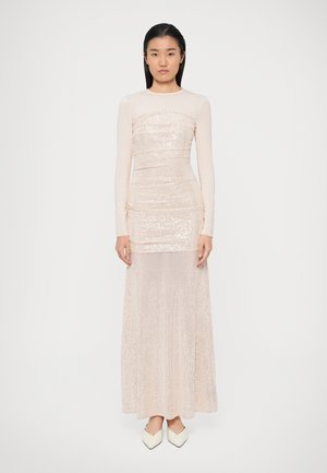 Sieviete, ģērbusies bālganrozā garajā piedurknē sekojošā, sešu apdara maxi kleitā un baltos, norādītiem apaviem, stāvot pret vienkāršu baltu fonu.