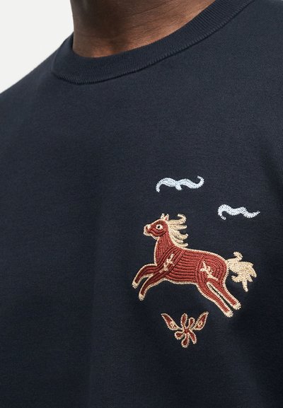 Μπλε πουκάμισο ναυτικού με κεντητό σχέδιο ενός καφέ αλόγου με κρέμ χαίτη, κρέμ ουρά, δύο μπλε σύννεφα από πάνω και ένα λουλουδένιο σχήμα κάτω.