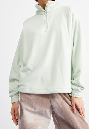 Persoon draagt een losse, licht mintgroene kwart-zip fleece trui en wijde beige broek, staand met één hand langs het lichaam.