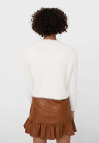 Eine lockige Person trägt große Ohrringe, einen weißen, flauschigen Pullover und einen Minirock aus braunem Leder mit Rüschensaum. Sie steht mit dem Rücken zu einem schlichten Hintergrund.
