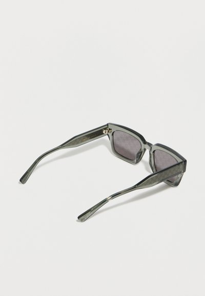 Ορθογώνια γυαλιά ηλίου με σκούρο φιμέ φακό, διαφανή γκρι πλαίσια και διακοσμητικά πλαϊνά, ακουμπισμένα σε λευκή επιφάνεια.