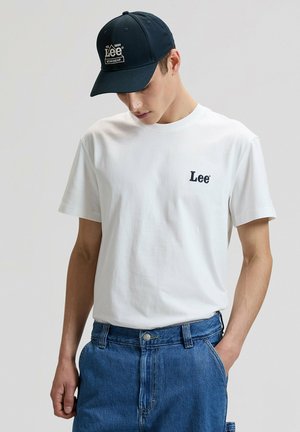 Biała bawełniana koszulka z krótkim rękawem, z niebieskim haftowanym logo "Lee" na lewej piersi. W zestawie z niebieskimi szortami dżinsowymi i granatową czapką.