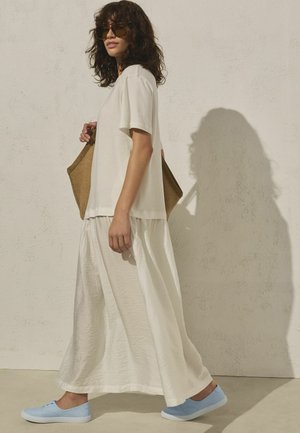 Femme aux cheveux bouclés portant des lunettes de soleil, une tenue blanche, des baskets bleu clair, et tenant un sac marron, marchant devant un mur texturé.