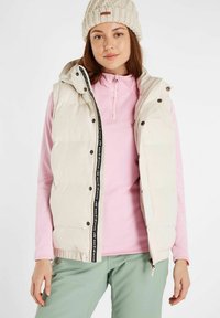 Gilet rembourré beige avec capuche et fermeture éclair à l'avant, orné d'un ruban noir avec logo. Porté par-dessus un haut à manches longues rose et associé à un pantalon vert.