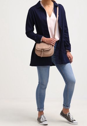 Vrouw die een donkerblauw jasje, witte blouse, lichtblauwe spijkerbroek, grijze sneakers draagt en een beige crossbodytas bij zich heeft, staand met één been licht gebogen.