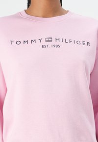Rožinė sportinė palaidinė, pagaminta iš švelnaus audinio, su apvalia apykakle ir spausdintu "TOMMY HILFIGER" logotipu, žemiau kurio yra užrašas "EST. 1985".