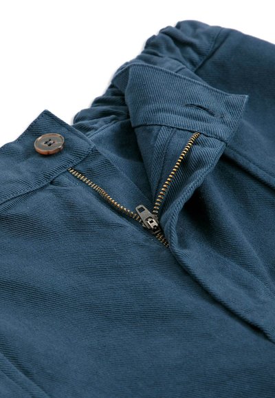 Gros plan d’un pantalon bleu marine avec une fermeture éclair en laiton partiellement ouverte et un bouton marron sur une ceinture en tissu avec dos élastique.