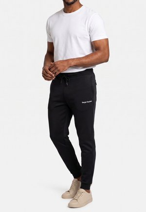 Hombre con camiseta blanca, pantalones jogger negros Sergio Tacchini y zapatillas beige, de pie con las manos juntas al frente.