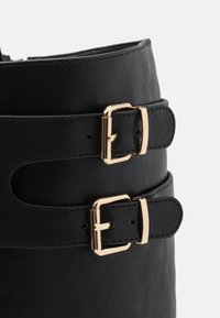 Black leather ankle boot with two gold buckle accents and stitched detailing along the edges. Smooth texture with a structured shape.
