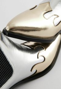 Botas tobilleras metálicas plateadas y doradas con puntas afiladas, acentos texturizados y diseños recortados en ondas, que presentan un panel negro elástico.