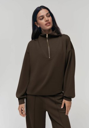 Žena s dlhými tmavými vlasmi, ktorá nosí hnedý pulóver na zips a zodpovedajúce nohavice, jemne sa pozerá doľava s neutrálnym pozadím.