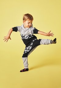 Czarny i szary kombinezon dziecięcy z wzorem graficznym, długimi rękawami i kapturem. Dziecko wykonuje dynamiczną pozę na żółtym tle.