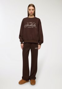 Set di felpa marrone e pantaloni abbinati, con logo stampato bianco "Stockholm Leisure Club". Tessuto morbido, vestibilità comoda e gambe larghe.