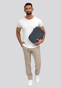 Borsa organizzativa grigia tenuta da una persona, forma rettangolare, materiale texturizzato, con un design semplice. Indossa una maglietta bianca e pantaloni beige.