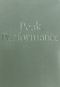 Sweat-shirt vert avec texte brodé "Peak Performance" et "Est. 1986" en vert plus clair. Texture lisse avec un col rond classique.