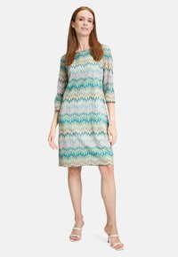 Kurzes Kleid mit kurzen Ärmeln in einem Zickzackmuster in den Farben Teal, Grün und Beige. Hergestellt aus leichtem Stoff mit glatter Textur.