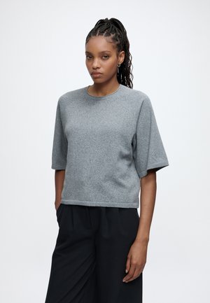 Young woman with braided hair wearing a grey short-sleeve sweater and black wide-leg pants standing against a plain background.