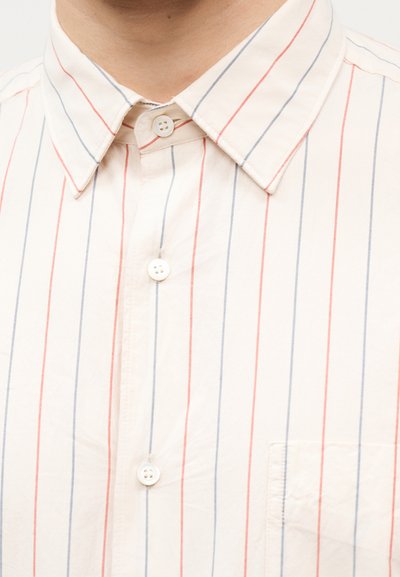 Λευκό πουκάμισο με κουμπιά, με κάθετες κόκκινες και μπλε ρίγες, γιακά με μυτερή άκρη και τσέπη στο στήθος στη δεξιά πλευρά.