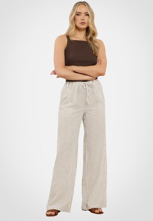 Femme blonde debout, les bras croisés, portant un haut marron sans manches et un pantalon blanc taille haute avec de fines rayures verticales noires.