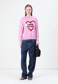 Maglione rosa con grafica a forma di cuore e testo "DAL FUTURO", abbinato a jeans bleu larghi, borsa nera e sandali. Design semplice e moderno.