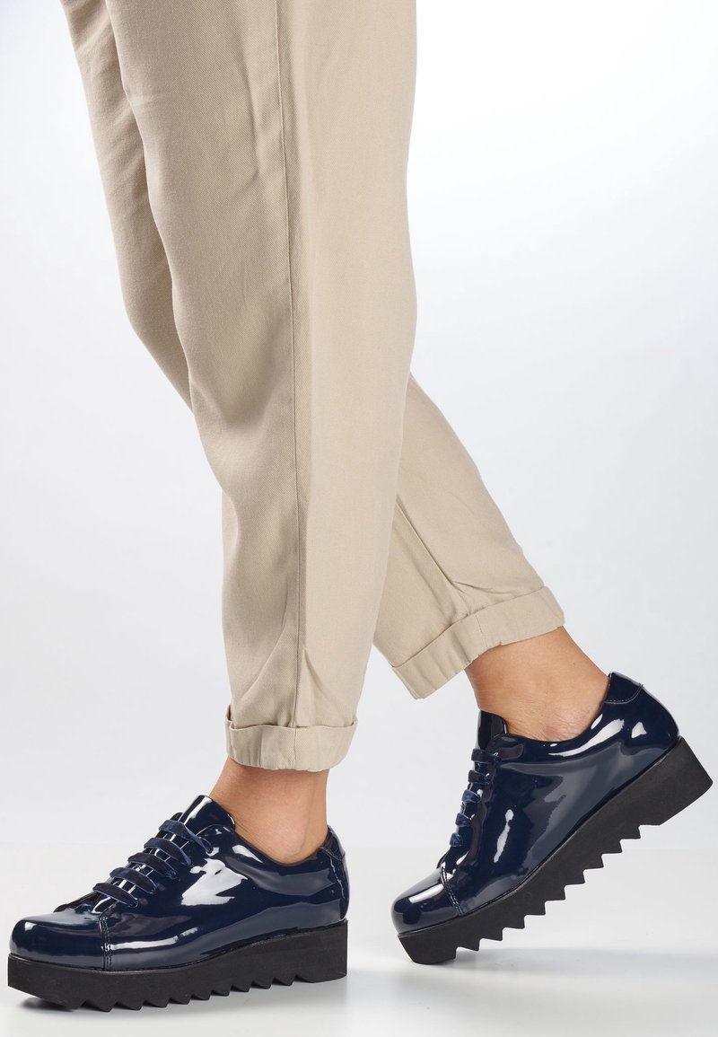 Henkilö, joka käyttää beigejä housuja ja kiiltäviä tummansinisia korkokenkiä, joissa on paksut, sahalaitaiset mustat pohjat, seisoo valkoista taustaa vasten.