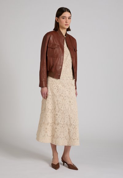 Kobieta w beżowej sukience midi w wzory i brązowej skórzanej kurtce, nosząca brązowe buty na niskim obcasie w szpic, stojąca na tle jednolitego szarego tła.