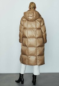 Cappotto lungo imbottito marrone con capuccio, caratterizzato da un design trapuntato e una texture lucida, indossato sopra pantaloni bianchi e stivaletti neri.