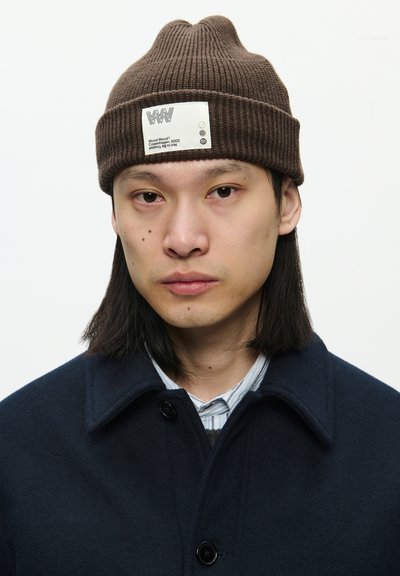 Rudas juostelėmis megzta kepurė su etiketė, kuriame yra logotipas ir tekstas. Dėvima su tamsiai mėlynu apykakle paltu ir dryžuota marškinėliais po juo.