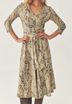 Femme portant une robe portefeuille longueur genou beige et marron à imprimé serpent, manches trois quarts et ceinture nouée à la taille.