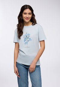 T-shirt di cotone azzurro con maniche corte, caratterizzato da un motivo floreale in tonalità di blu e viola, e la scritta "Le Jardin Parisien."
