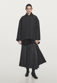 Black quilted jacket with a collar, paired with a long black skirt. The outfit features pockets and a smooth texture.  Black ankle boots complete the look.
